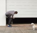 A man in a plaid shirt manually lifts a white garage door while a small white dog stands nearby on a brick driveway.