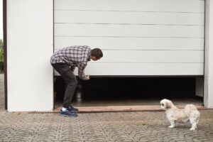 A man in a plaid shirt manually lifts a white garage door while a small white dog stands nearby on a brick driveway.