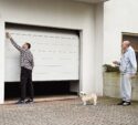 A man in a plaid shirt opens a garage door while another man in gray sweatpants stands nearby holding a remote, with a small dog beside him.