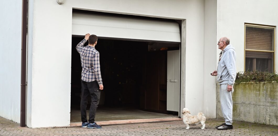 Two men and a small dog stand outside an open garage door attached to a white building. One man is facing the garage, and the other is standing nearby with the dog.