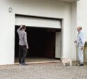 Two men and a small dog stand outside an open garage door attached to a white building. One man is facing the garage, and the other is standing nearby with the dog.