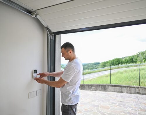 A man in a white t-shirt enters a code on a wall-mounted keypad inside a garage with the door open, overlooking a paved driveway and greenery outside.