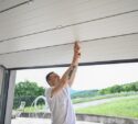 A person in a white shirt adjusts or repairs the inside of a partially open garage door during the daytime.