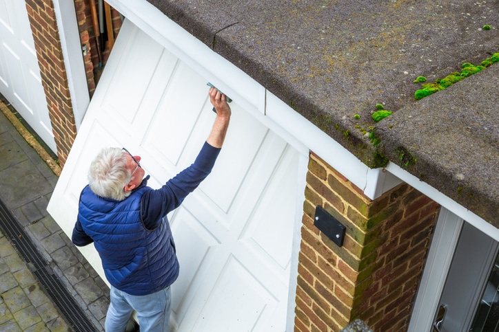 An older man with gray hair is cleaning or repairing a white garage door attached to a brick house, with moss visible on the roof edge.