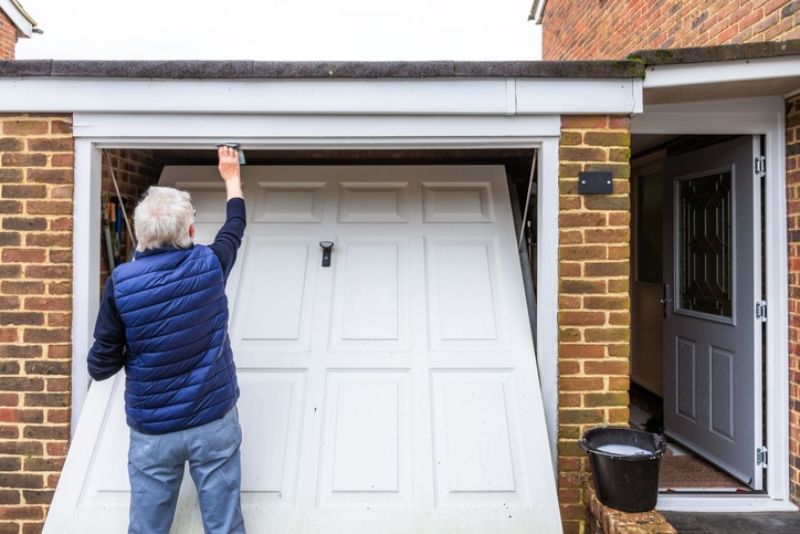 An older person in a blue vest wipes the top of a white garage door that is partially open, with a bucket nearby on the ground.