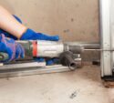 A person wearing blue gloves uses a power drill to bore a hole into a metal frame at a construction site.