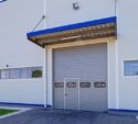 Gray industrial garage door with small windows in a white commercial building, surrounded by pavement and green grass.