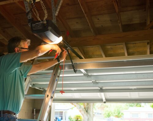 A man standing on a ladder repairs a garage door opener in a residential garage with the door partially open.