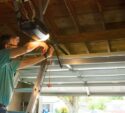 A man stands on a ladder adjusting a garage door opener inside a partially open garage with a wooden ceiling.