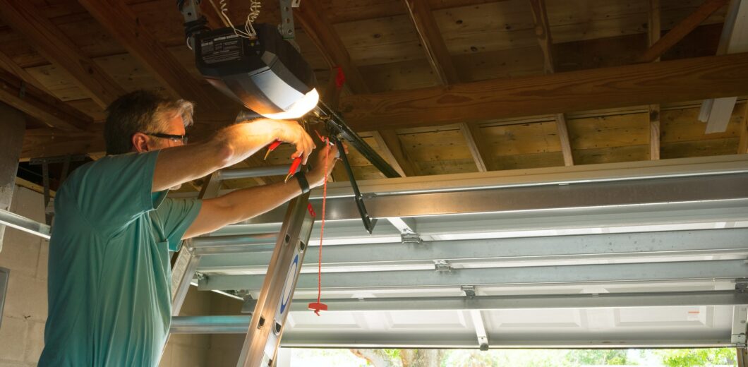 A man standing on a ladder repairs an automatic garage door opener inside a garage with the door partially open.