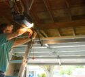 A man standing on a ladder repairs an automatic garage door opener inside a garage with the door partially open.