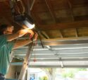A person stands on a ladder adjusting a garage door opener mechanism inside a partially open garage.