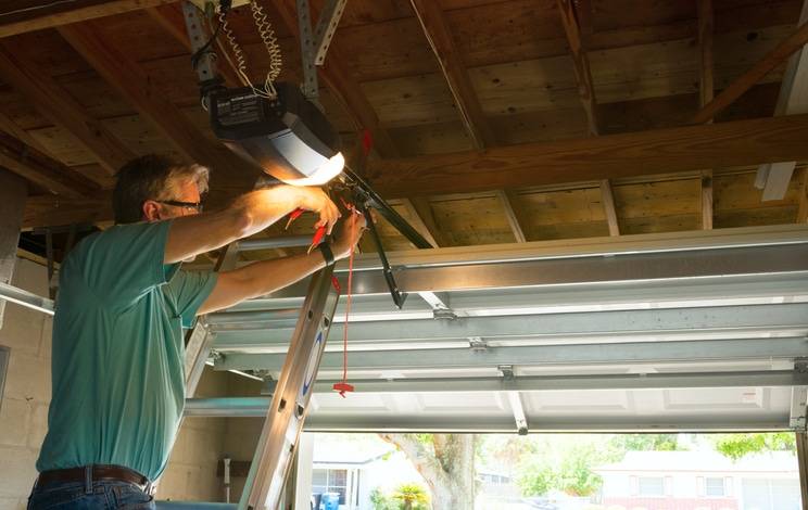A man stands on a ladder adjusting a garage door opener inside a partially open garage with a wooden ceiling.