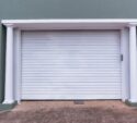 A closed white garage door with vertical ridges, framed by two white columns against a green wall. Concrete driveway in front.