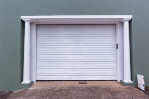 A closed white garage door with vertical ridges, framed by two white columns against a green wall. Concrete driveway in front.