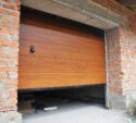 Partially open wooden garage door on a brick garage under construction, with building materials scattered on the unfinished concrete floor.
