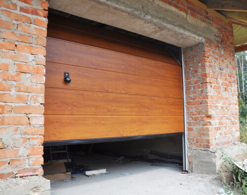 Partially open wooden garage door on a brick garage under construction, with building materials scattered on the unfinished concrete floor.