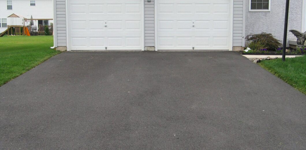 A double garage with two white doors and a freshly paved asphalt driveway in front of a light gray house.