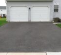 A double garage with two white doors and a freshly paved asphalt driveway in front of a light gray house.