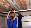 Two workers in blue uniforms install an automatic garage door opener on the ceiling of a garage with an unfinished wooden roof.