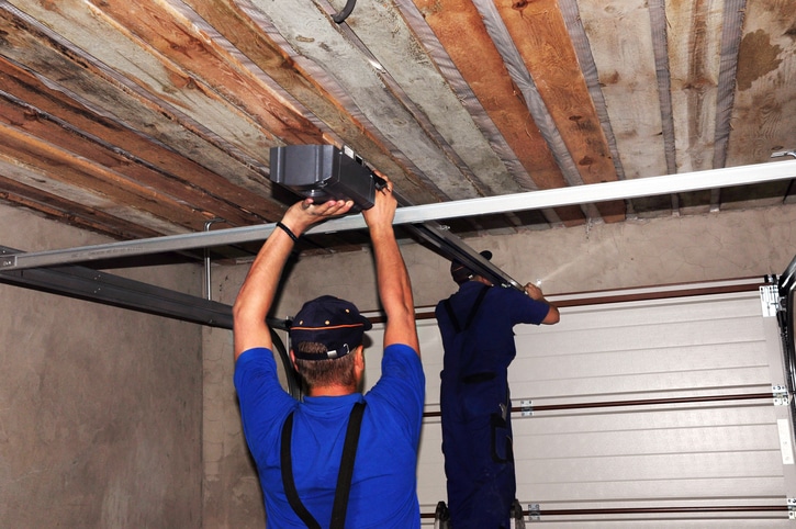 Two workers in blue uniforms install an automatic garage door opener on the ceiling of a garage with an unfinished wooden roof.