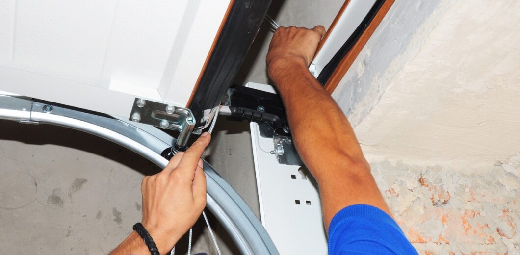 Person installing or repairing a garage door mechanism, using tools to adjust components near the door’s top corner.