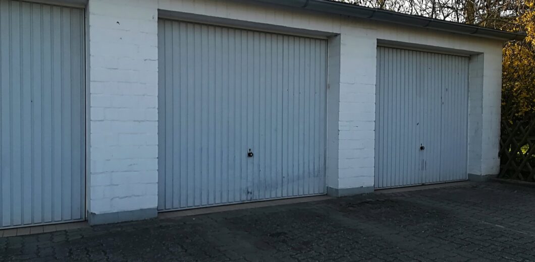 Three closed, white, single-car garage doors with vertical panels are set in a white brick building on a paved driveway.