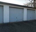 Three closed, white, single-car garage doors with vertical panels are set in a white brick building on a paved driveway.