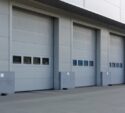 Four closed industrial garage doors on a gray building, each with a row of rectangular windows and protective barriers at the base.