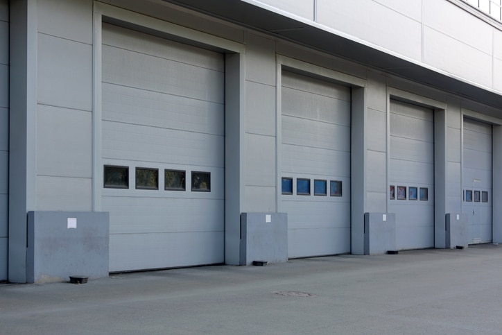 Four closed industrial garage doors on a gray building, each with a row of rectangular windows and protective barriers at the base.