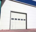 Large white industrial garage door with five horizontal windows and a small pedestrian door at the bottom, set in a warehouse exterior with blue trim and red brick pavement.
